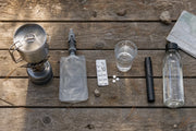 Vier manieren om water te zuiveren op een picknicktafel: kookpannetje met brander, waterfilter, zuiveringstabletten en UV-C-pen